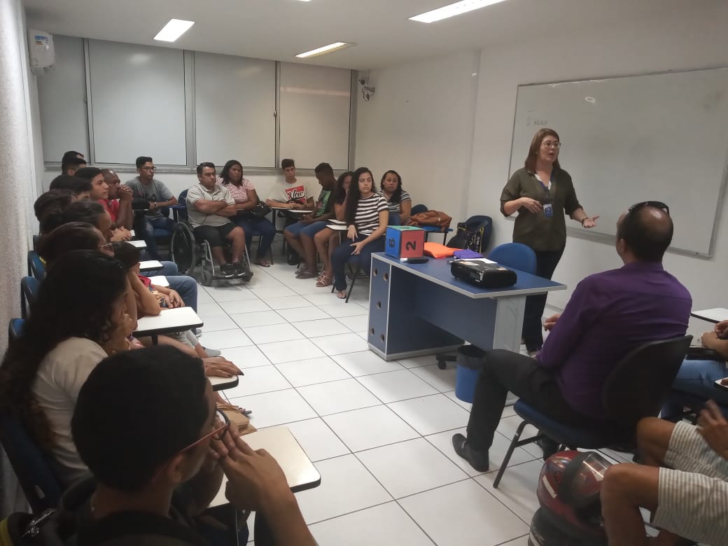 pessoas sentadas em carteiras em sala de aula e professora falando em pé no meio